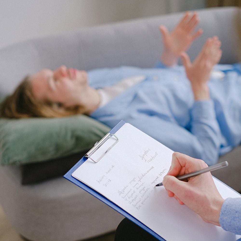 Patient lying on a couch with psychologist taking notes as they speak.